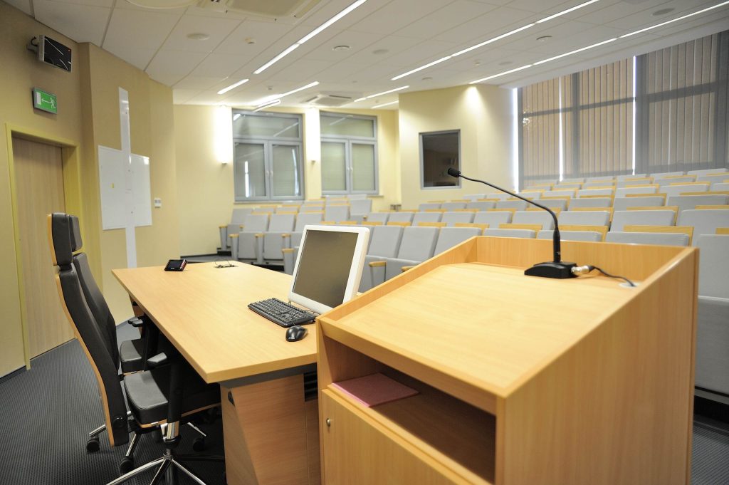 Sala wykładowa z rzędami miejsc, mównicą, mikrofonem i stanowiskiem komputerowym.