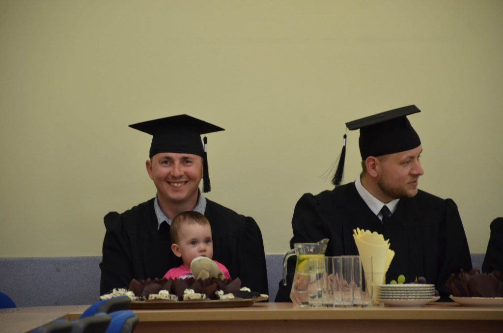 Dwaj absolwenci w togach i biretach siedzą przy stole; jeden trzyma małe dziecko.