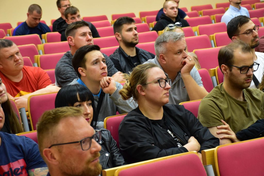 Uczestnicy wykładu siedzą w czerwonych fotelach, spoglądając w stronę prowadzącego.