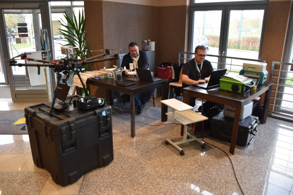 Stanowisko pokazowe z dronami, aparaturą i komputerami w holu podczas konferencji naukowej.