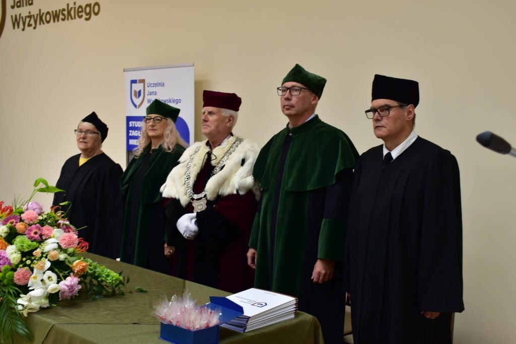 Przedstawiciele uczelni w togach stoją przy stole z kwiatami podczas zakończenia roku PUTW.