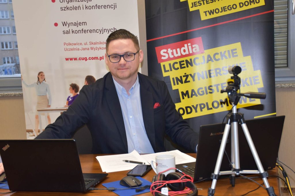 Mężczyzna przy biurku z laptopami i kamerą podczas spotkania na uczelni.