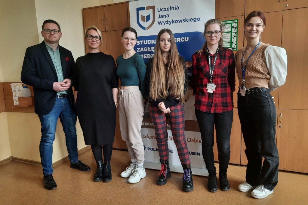 5 kobiet i 1 mężczyzna stoi na tle rollupa z reklamą uczelni