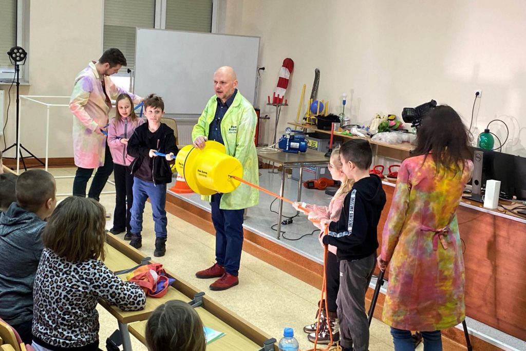 Dzieci uczestniczą w pokazie doświadczenia z żółtym urządzeniem w sali laboratoryjnej.
