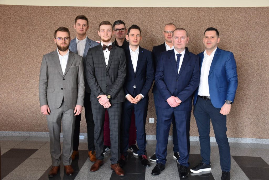 Grupa mężczyzn w strojach oficjalnych pozuje w holu uczelni.