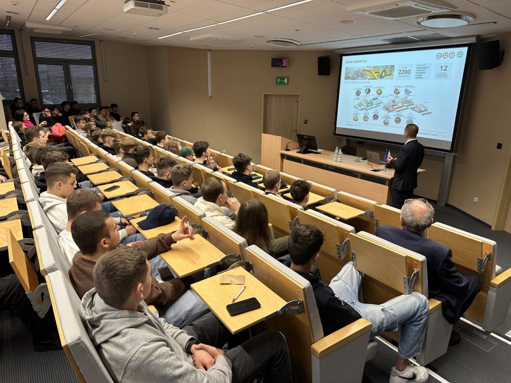 Prowadzący prezentuje slajd publiczności w auli podczas Dnia Robotyki.