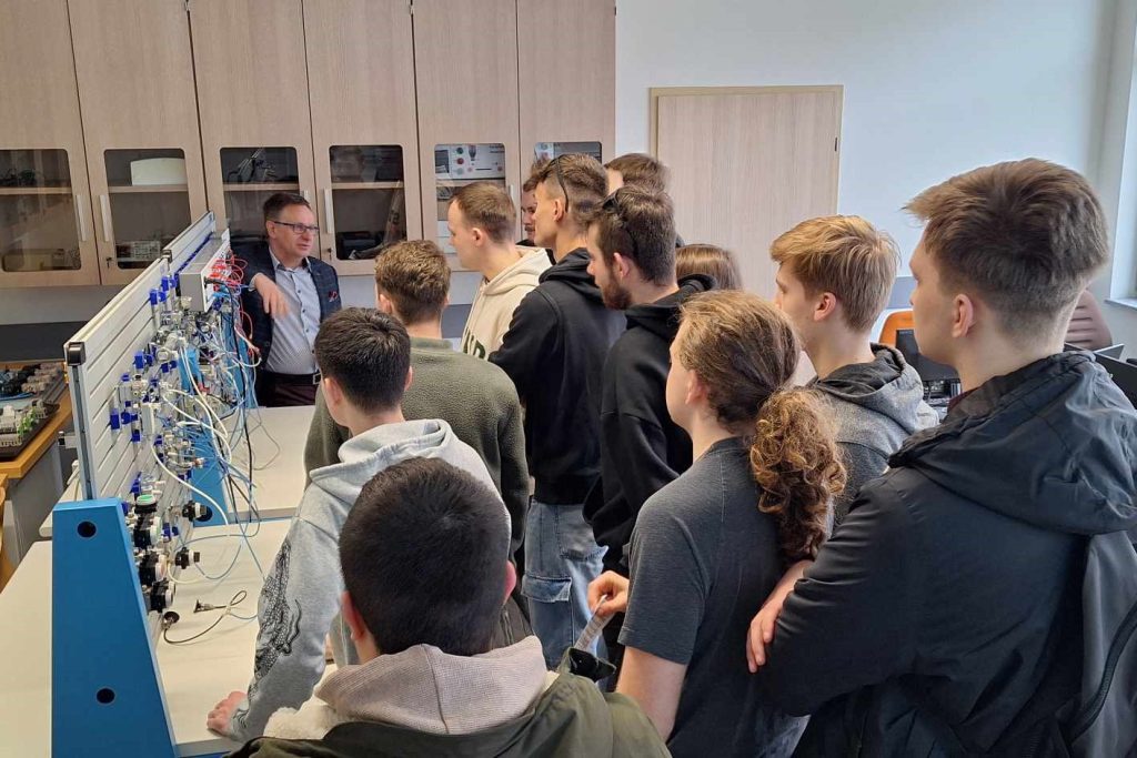Grupa uczniów obserwuje pokaz przy stanowisku laboratoryjnym z układami pneumatycznymi.