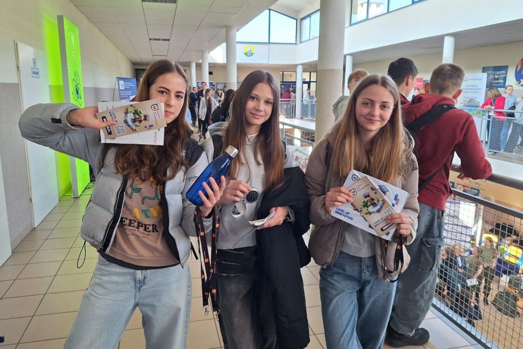 Trzy uczestniczki wydarzenia prezentują materiały informacyjne w holu szkoły.