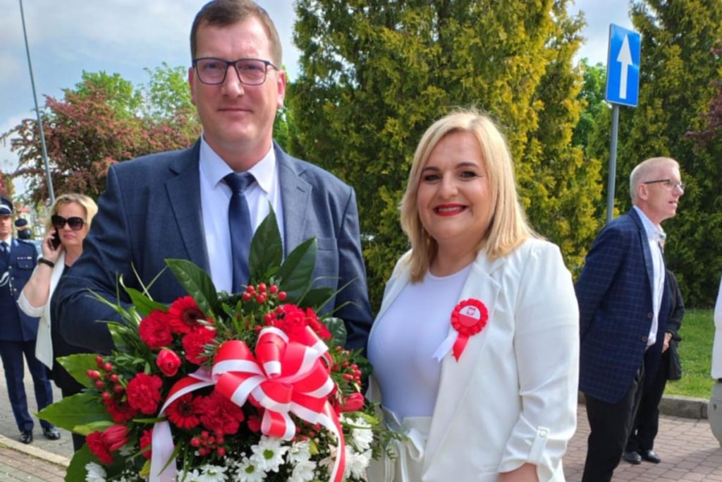 Kobieta i mężczyzna prezentują wieniec z biało-czerwoną wstęgą podczas uroczystości plenerowej.