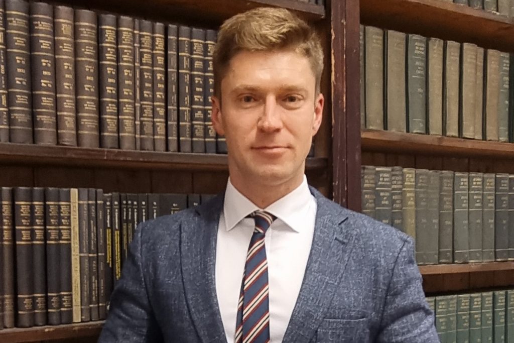 Portret mężczyzny w garniturze na tle regału z książkami.