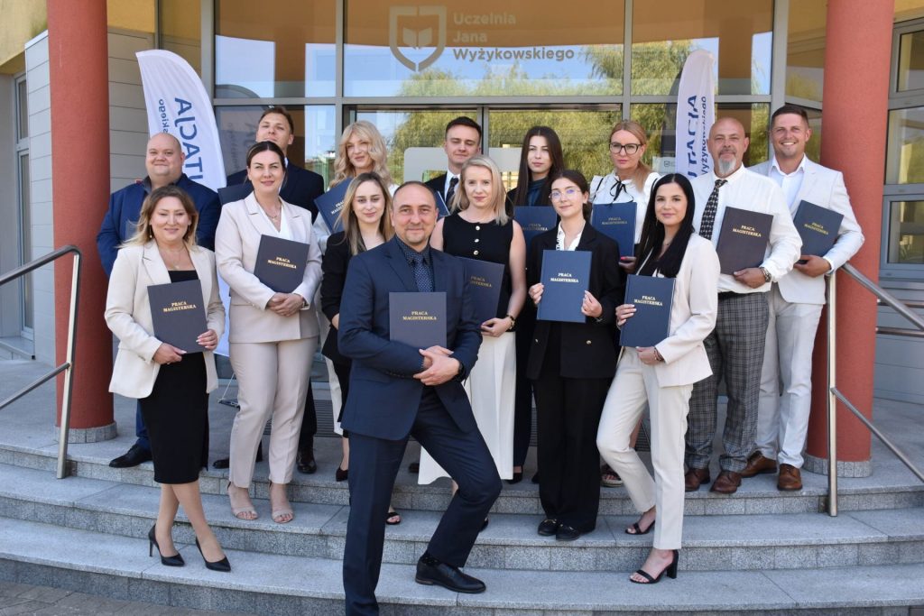 Absolwenci prezentują oprawione prace na schodach przed UJW; mężczyzna z przodu pozuje z szeroko rozstawionymi nogami.