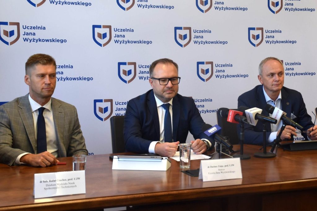 Trzej przedstawiciele uczelni siedzą przy mikrofonach podczas konferencji prasowej.