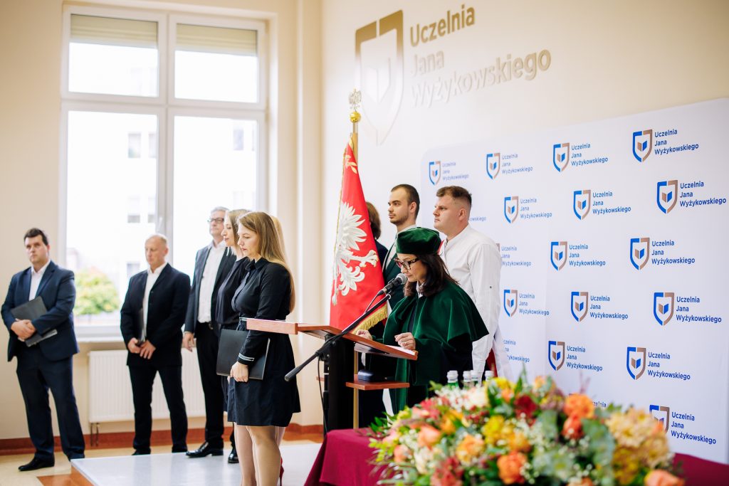 Uczestnicy inauguracji stoją w auli, a osoba w stroju akademickim przemawia przy mównicy.