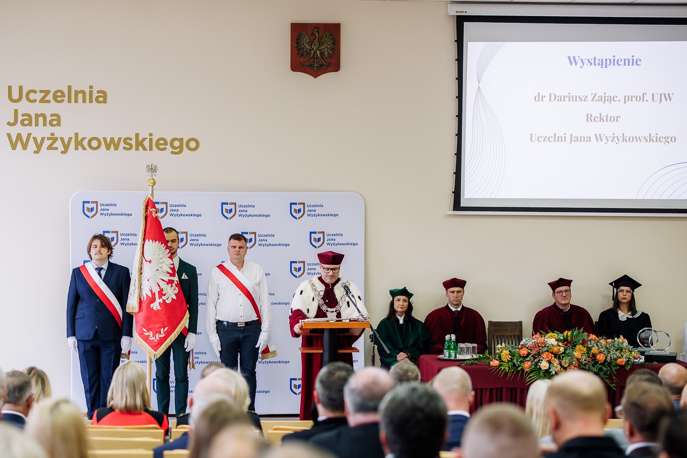 inauguracja nowego roku akademickiego. Na zdjęciu są władze uczelni, sztandar UJW, zaproszeni goście, w tle wyświetla sie napis - wystapienie rektora UJW dra Dariusza Zająca