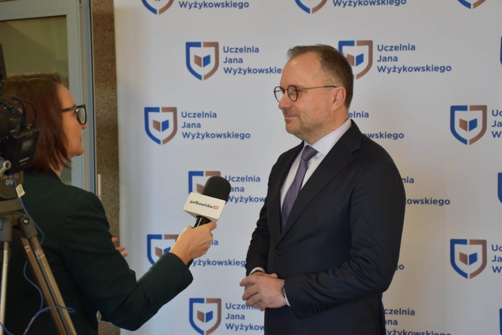 Mężczyzna udziela wywiadu dziennikarce z mikrofonem na tle logo UJW.