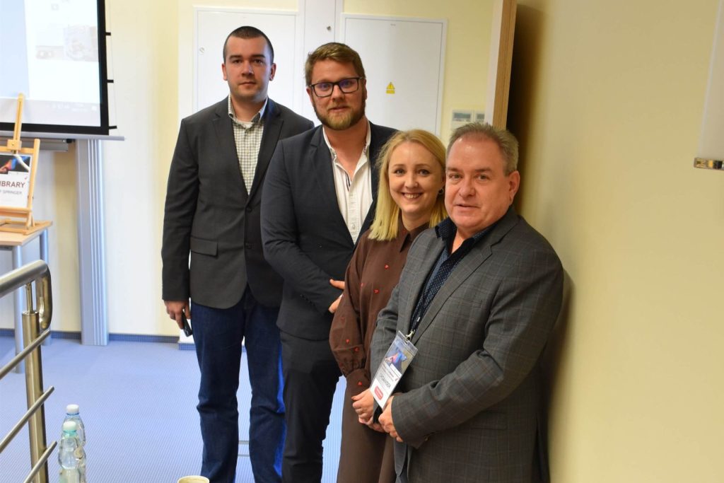 Czworo uczestników konferencji dotyczącej zarządzania danymi pozuje w sali wykładowej.