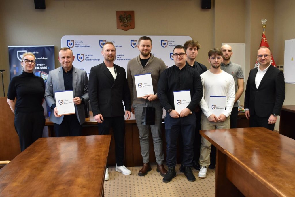 Uczestnicy uroczystości prezentują teczki z dokumentami na tle planszy UJW.