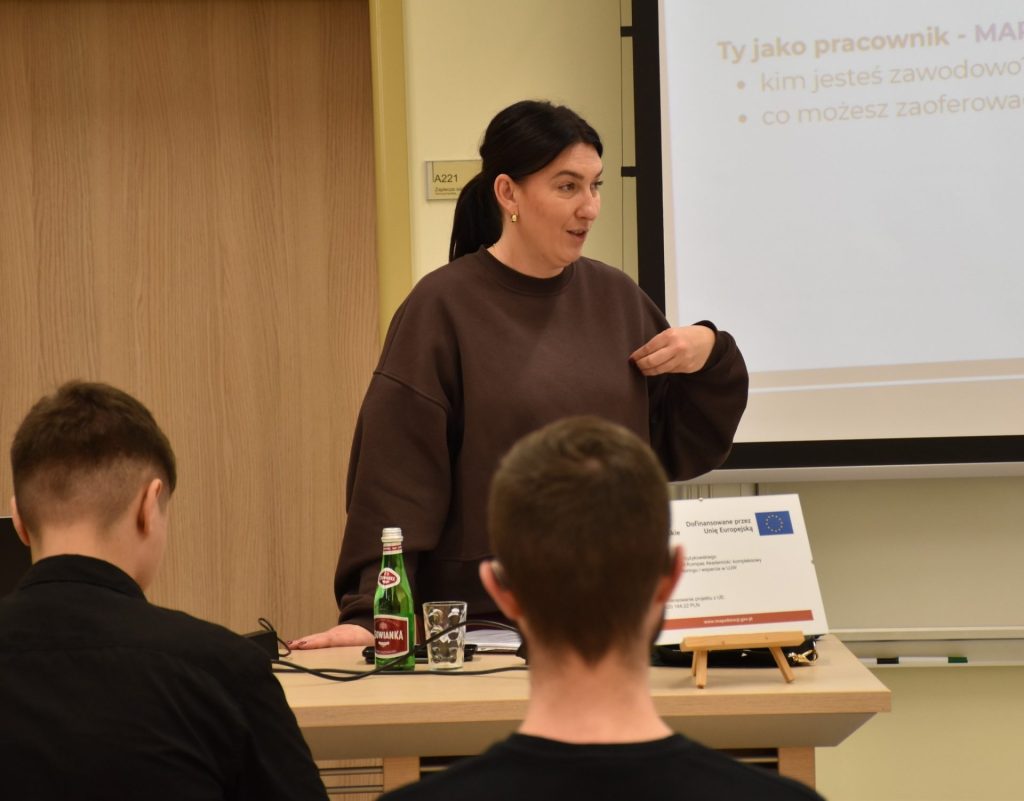 Prowadząca warsztaty dotyczące CV przemawia przy ekranie do uczestników siedzących przy stołach.