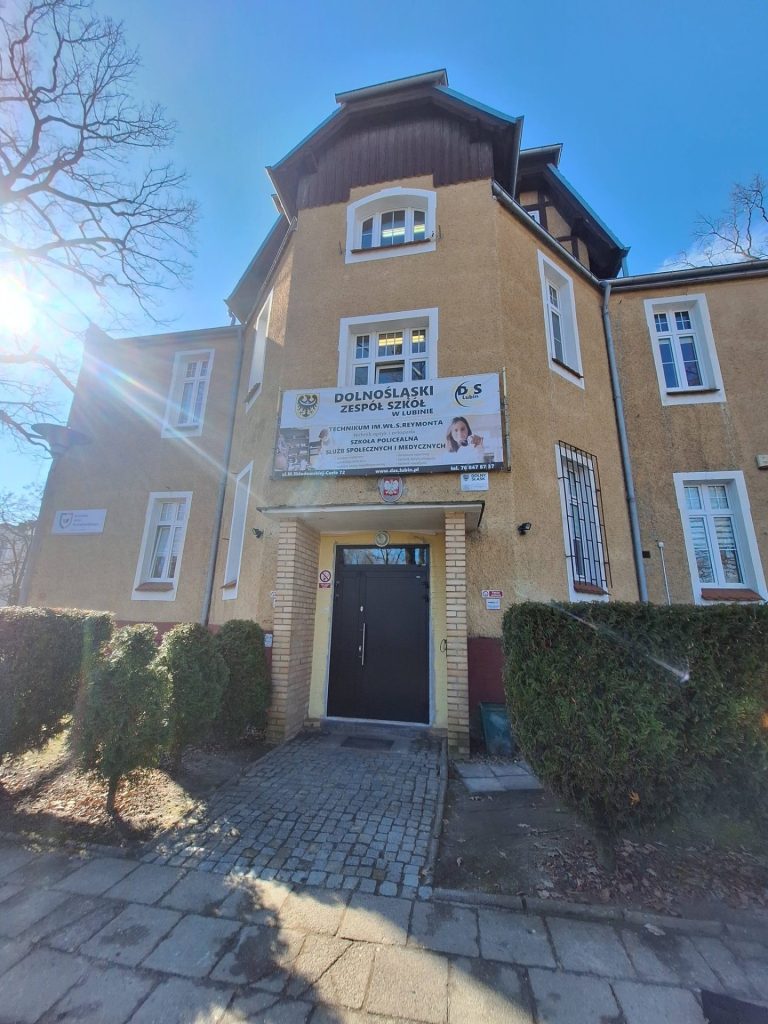 Front ceglanego budynku szkoły z wejściem i wieżyczką, odwiedzanej przez przedstawicieli UJW.