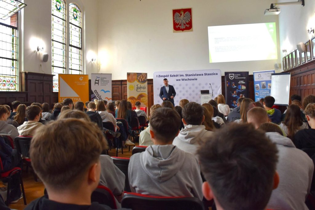 Uczniowie słuchają wystąpienia w szkolnej auli przy stoiskach informacyjnych.