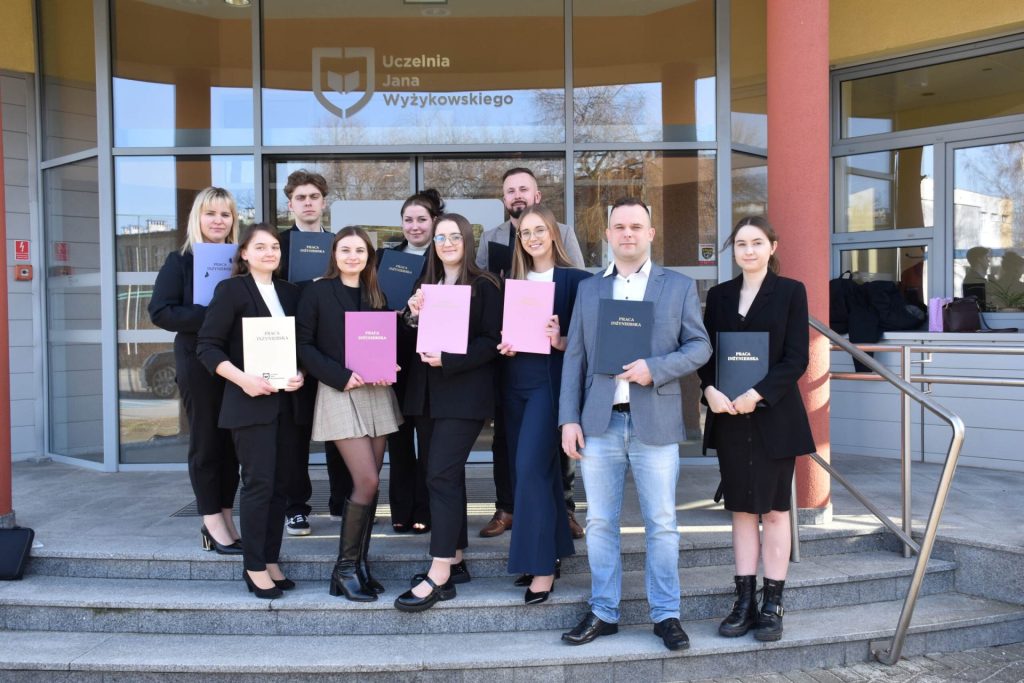 Absolwenci z oprawionymi pracami dyplomowymi pozują na schodach przed wejściem do UJW.