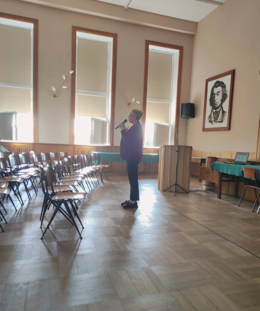 Prowadząca stoi między rzędami krzeseł w szkolnej auli.