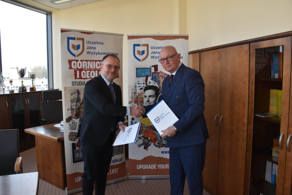 Dwaj mężczyźni podają sobie dłonie i prezentują dokumenty przy planszach UJW.