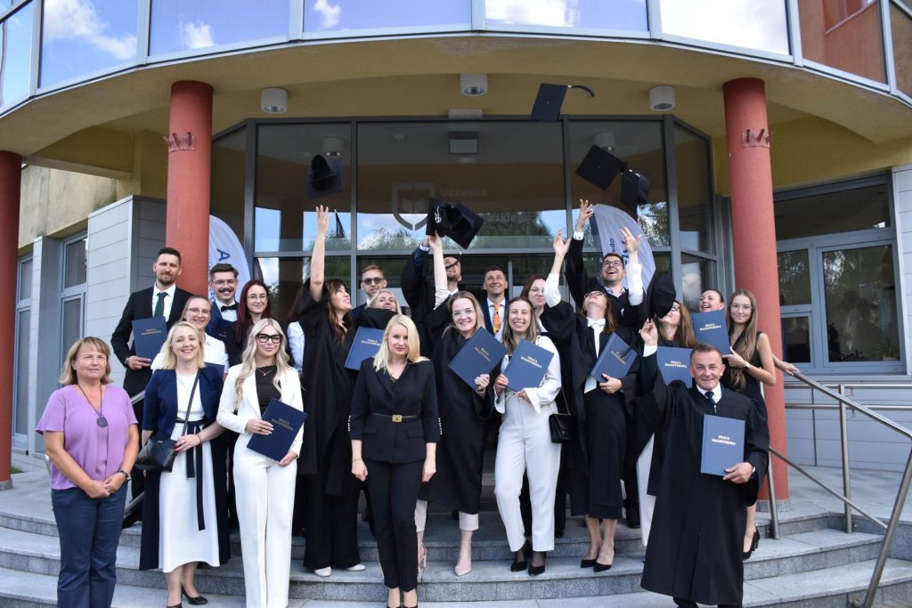 Grupa absolwentów i uczestników uroczystości przed wejściem do uczelni; absolwenci unoszą birety.