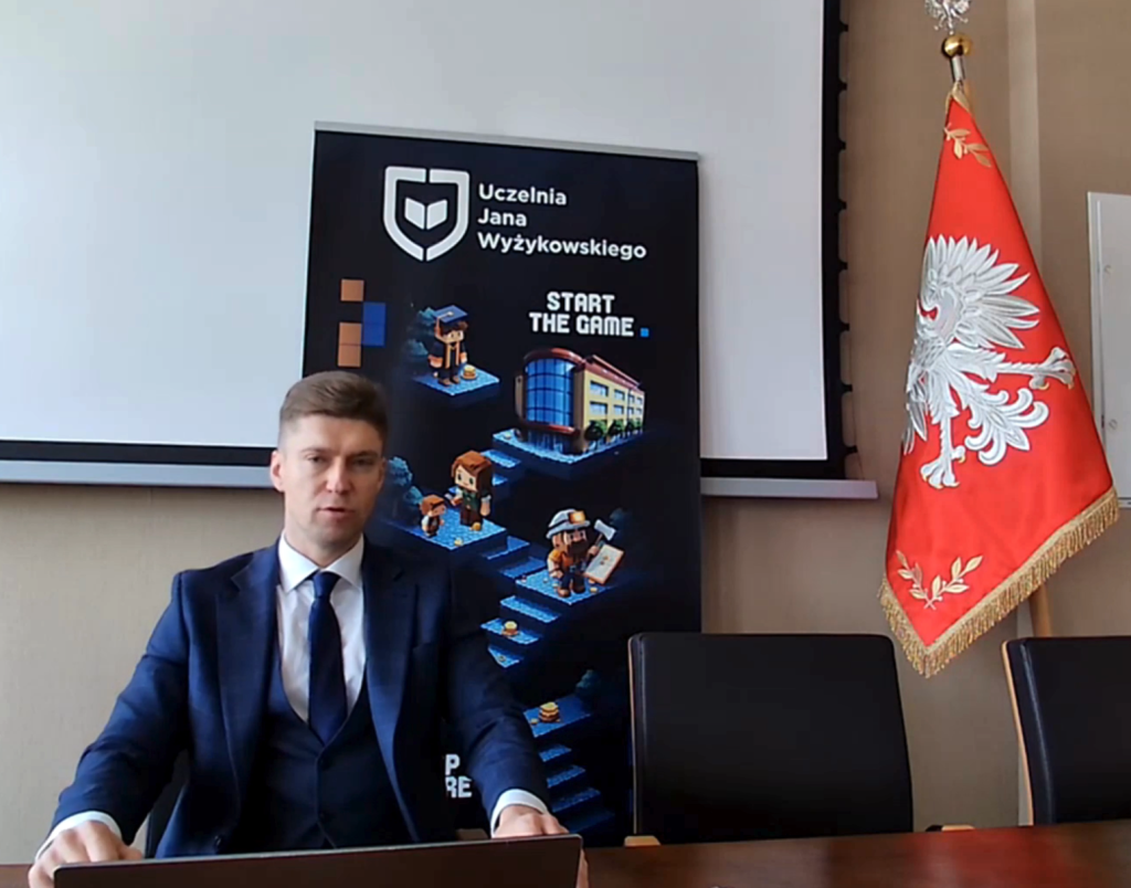 Mężczyzna w garniturze siedzi przy stole na tle planszy UJW i flagi Polski.