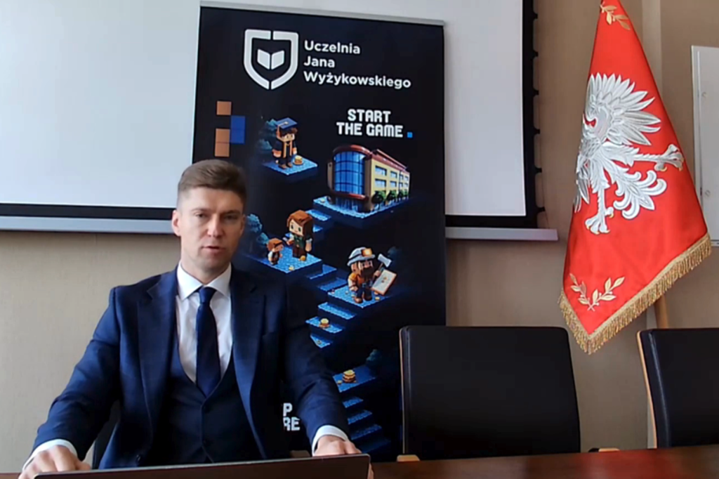 Mężczyzna w garniturze siedzi za biurkiem przy planszy UJW i fladze z polskim godłem.