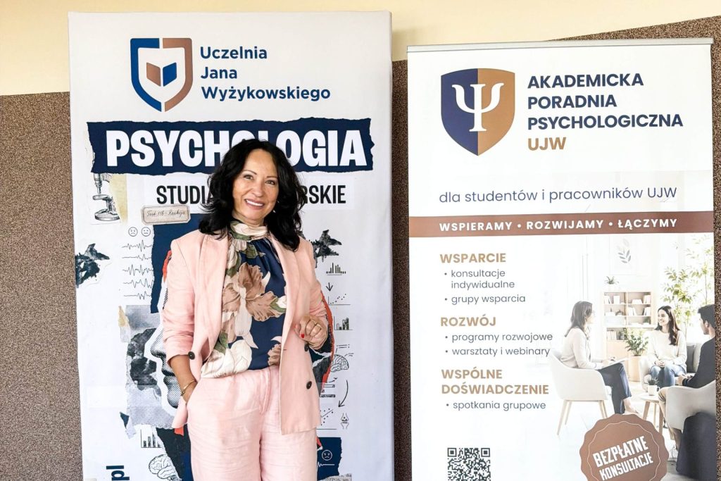 Kobieta przemawia z mikrofonem przy planszach Akademickiej Poradni Psychologicznej i kierunku Psychologia UJW.