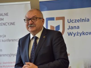 Mówca w okularach i granatowym garniturze na tle banerów UJW.
