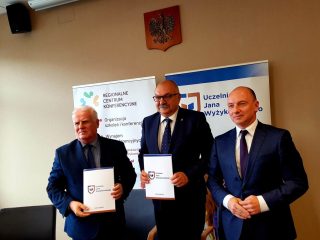 Trzej przedstawiciele pozują z dokumentami na tle banerów UJW.