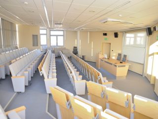 Pusta sala wykładowa z rzędami foteli na podestach i biurkiem prowadzącego.