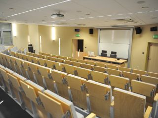 Amfiteatralna sala UJW z pulpitami i stołem prezydialnym pod ekranem.