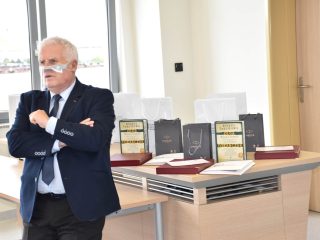 Siwowłosy mężczyzna w garniturze i osłonie twarzy stoi przy stole z książkami, dyplomami i torbami.