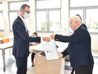 Siwowłosy mężczyzna przekazuje białą torbę upominkową mężczyźnie w okularach.