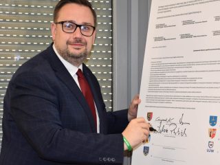 Mężczyzna w okularach i czerwonym krawacie pozuje przy podpisanym porozumieniu.