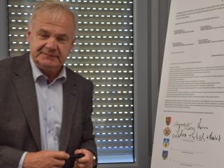 Siwowłosy uczestnik w szarej marynarce stoi przy planszy porozumienia.