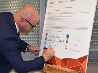 Mężczyzna w okularach podpisuje porozumienie klastra na dużej planszy.
