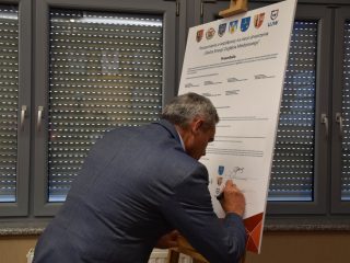 Siwowłosy mężczyzna w szarej marynarce składa podpis na porozumieniu.