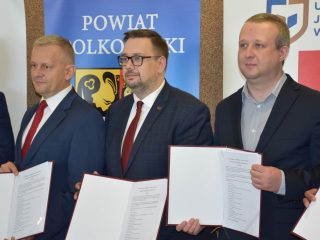 Trzej sygnatariusze prezentują dokumenty na tle banera Powiatu Polkowickiego.