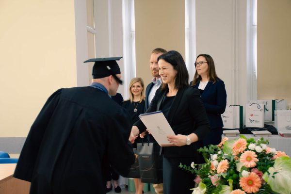 Kobieta ściska dłoń i wręcza teczkę męźczyźnie, który ubrany jest w czarną togę i biret