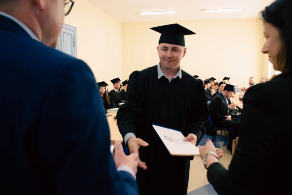 Mężczyzna ściska dłoń mężczyzny i wręcza mu teczkę. Mężczyzna ubrany jest w czarną togę i biret