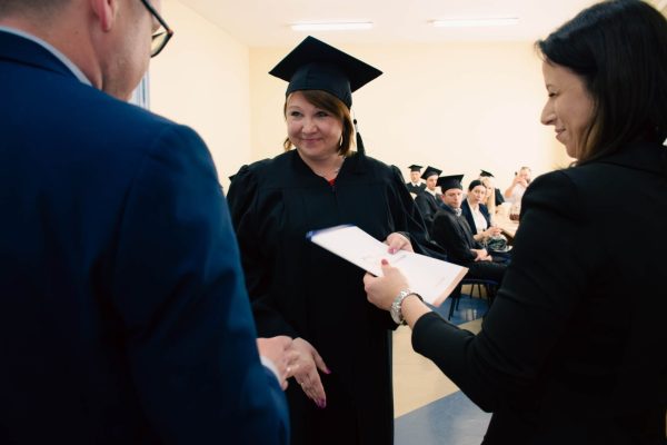 Mężczyzna ściska dłoń kobiety i wręcza jej teczkę. Kobieta ubrana jest w czarną togę i biret