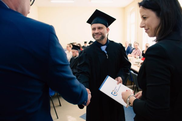 Mężczyzna ściska dłoń mężczyzny i wręcza mu teczkę. Mężczyzna ubrany jest w czarną togę i biret