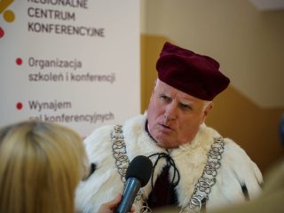 Rektor w uroczystym stroju i birecie udziela wywiadu reporterce z mikrofonem.