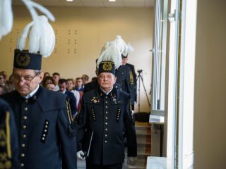 Chórzyści w mundurach górniczych i czakach z białymi pióropuszami przechodzą między rzędami foteli.