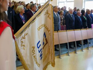 Sztandar uczelni na pierwszym planie, w tle stoją uczestnicy inauguracji roku akademickiego.