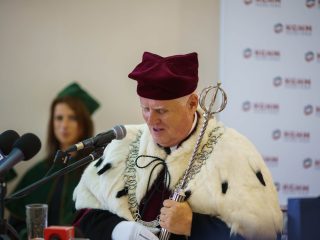 Rektor w uroczystym stroju z łańcuchem i berłem przemawia do mikrofonu.
